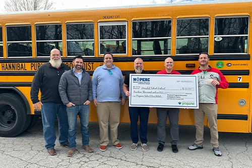 A group of smiling people hold a giant check in front of a school bus