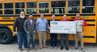 A group of smiling people hold a giant check in front of a school bus