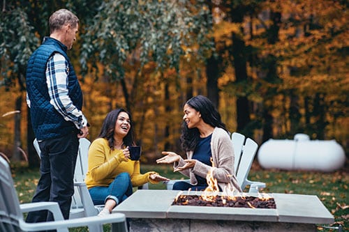 People conversing around a fire pit in front of a propane tank on a fall day
