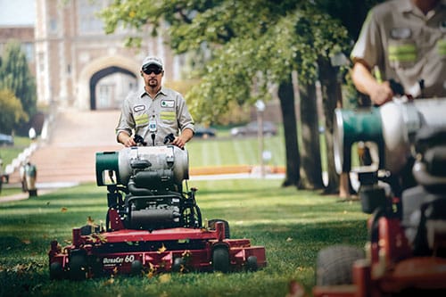 Two landscape contractors on stand on propane lawnmowers moving the grass on a college campus