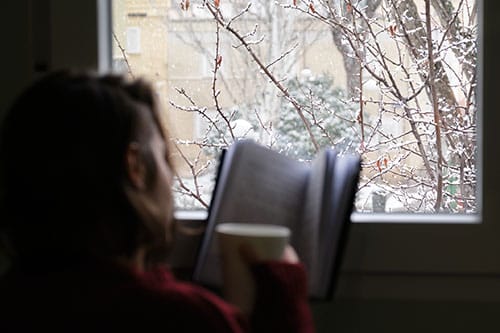 A woman reading and holding a cup of coffee in front of a window on a snowy day